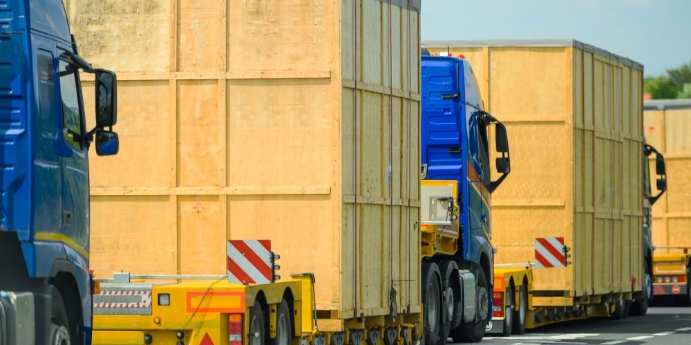 Heavy Oversize Load Convoy on the Route to Destination. Few Semi Trucks Pulling Large Wooden Oversized Containers. Transportation Industry.